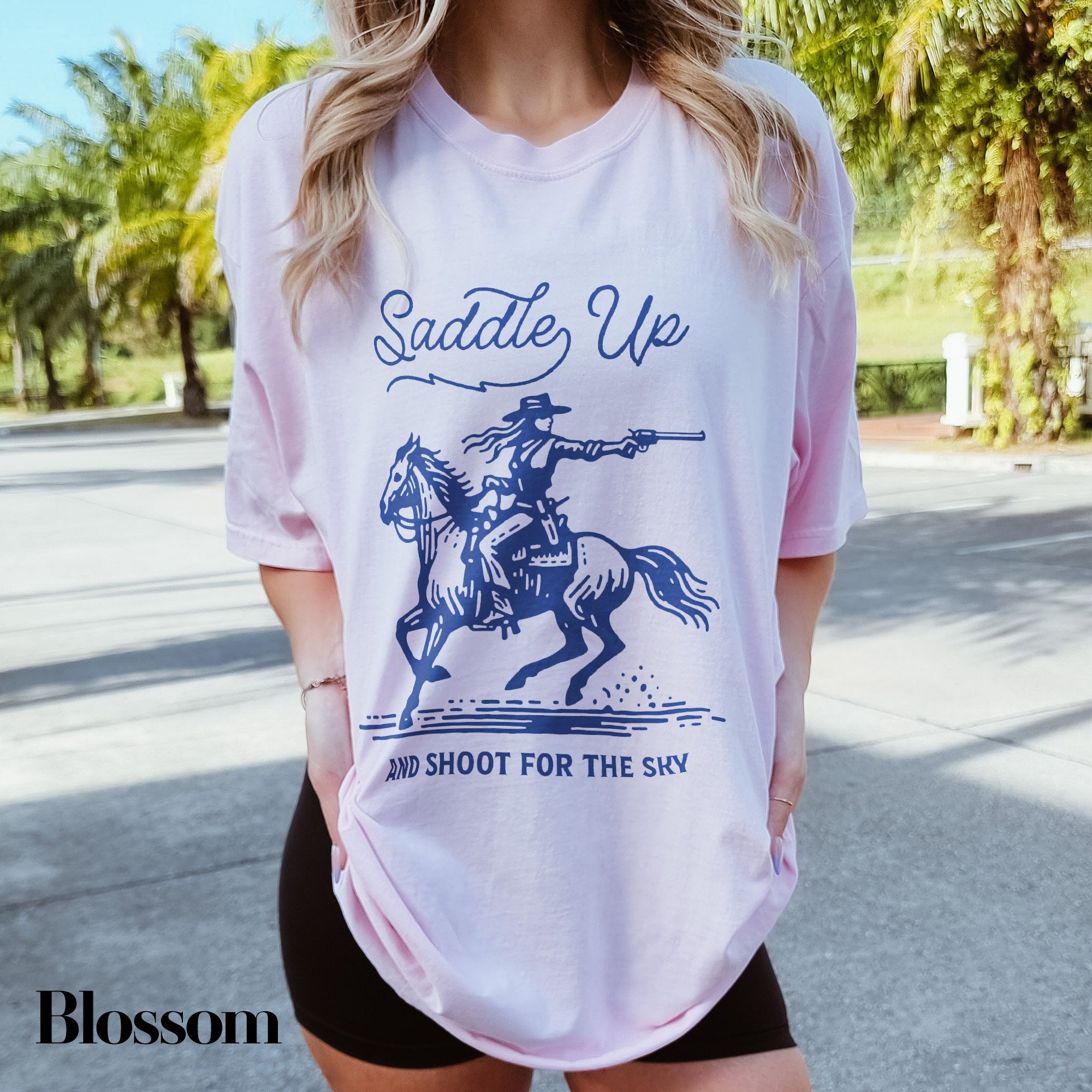 a woman wearing a t - shirt that says saddle up and shoot for the sky